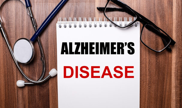 ALZHEIMERS DISEASE is written on white paper on a wooden background near a stethoscope and black-framed glasses. Medical concept