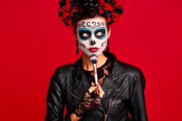 Girl apply facial makeup sugar skullby brush with a wreath of flowers on her head and skull, wearth lace gloves and leather jacket, isolated on red. concept of Halloween or La Calavera Catrina