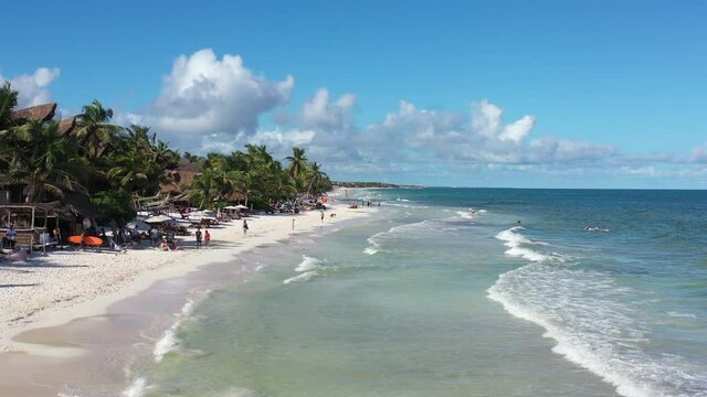 Vista a&eacute;rea sobre la playa y zona hotelera en Tulum Beach, con algunas personas caminando sobre la playa y un cielo azul y nublado como fondo.