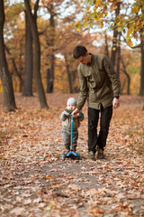 Fototapeta premium dad rolls a little son on a scooter in an autumn park. weekend spent with family. Concept of friendly family. 