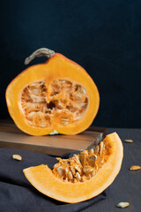 Ugly raw halved orange pumpkin laying on cutting board on brown textile on dark wooden table against black background representing autumn harvesting season and halloween holiday. Vertical orientation