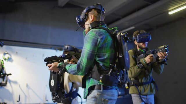 Medium shot of team of three young people, two men and woman, in virtual reality headsets and other gear playing VR adventure game. Friends shooting with gun controllers in defense
