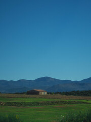 Casa en medio del campo con montañas en el fondo y un cielo azul despejado.