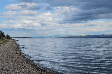 Fototapeta na wymiar Pebbles shore of clear calm undulating blue Lake Baikal, mountains on the horizon, clouds