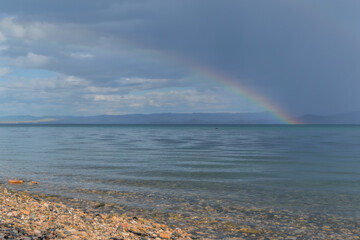 Colorful rainbow after rain above clear calm undulating blue water of Lake Baikal, pebbles shore, mountains on horizon. Landscape