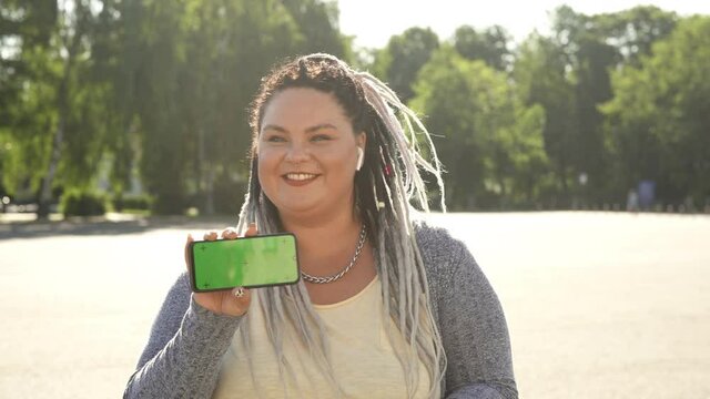 Commercial, copy space concept. Young pretty fat woman with dreads showing green screen phone to camera and dancing. Beautiful stout girl with chromakey on her smartphone advertising mobile app.