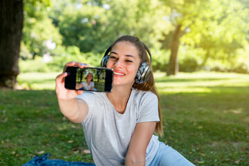 Young beautiful happy smiling woman taking selfie with her smartphone in the park while she listening the music over wireless headphones