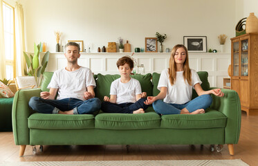 Calm young family with son practice yoga together sitting in Lotus position on couch. Conscious parents with school boy child rest on sofa meditating relieve negative emotions on weekend at home.