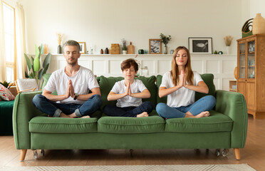 Serene couple and little son sitting on sofa in lotus position with closed eyes doing namaste...