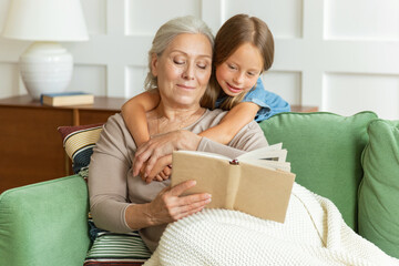 Pretty grandmother and little granddaughter reading book, sitting on comfortable couch in living...