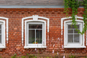 Fototapeta premium Old city architecture, red brick building with windows and plant hanging from roof. Vintage house in countryside. Retro dwelling in village or rural area. Abandoned construction or summer cottage