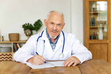 Smiling old male doctor in white uniform at desk fill patient form with medical history or writes patient prescription. Mature senior man GP physician makes notes in journal of client anamnesis.