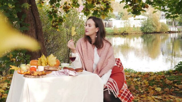Beautiful woman sitting at the table near the pond in the autumn park. Cozy weekend picnic with red wine and food, candles and pumpkin, colorful fall leaves