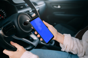 The girl in the car holds the steering wheel of the car. Girl looks at the smartphone screen. Blue...