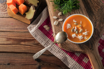  Pumpkin cream soup with cheese and seeds on a wooden table on a red checkered tablecloth. Nearby are the ingredients for cooking