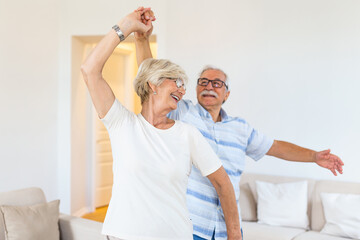 Carefree happy active old senior couple dancing jumping laughing in living room, cheerful retired elder husband holding hand of mature middle aged wife enjoy fun leisure retirement lifestyle at home