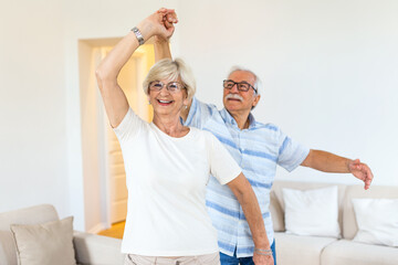 Obraz na płótnie Canvas Joyful active old retired romantic couple dancing laughing in living room, happy middle aged wife and elder husband having fun at home, smiling senior family grandparents relaxing bonding together