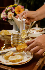 Obraz na płótnie Canvas Bee honey in glass container and hands holding honeydew, placing honey on toasted bread, on a table set with vintage style dishes and jug with blurred flowers in the background.