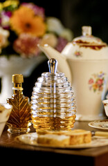 Bee honey in glass container and hands holding honeydew, placing honey on toasted bread, on a table set with vintage style dishes and jug with blurred flowers in the background.