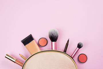 Overhead view of a beige leather make up bag, with cosmetic beauty products spilling out on to a pastel pink background