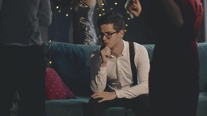 Sad and unhappy guy feeling lonely at celebrate at home, sitting on couch or sofa alone in front of having rest and fun people or friends. Portrait of upset man in decorated house at xmas party.