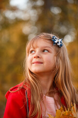 Fototapeta premium Outdoor portrait of happy blonde child girl in a red jacket holding yellow leaves. Little girl walking in the autumn park or forest. Warm sunny weather. Fall concept