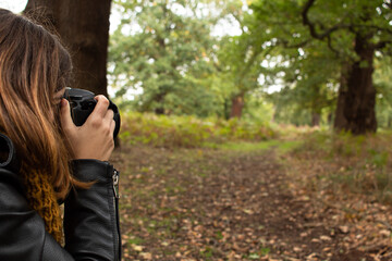 Attractive young female with a black leather jacket taking a photo with her reflex camera while walking on the park during autumn