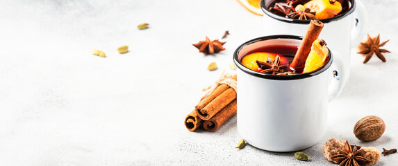 Mulled wine in white metal mugs with cinnamon, spices and orange on gray background, traditional...