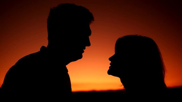 Closeup of silhouettes of man and woman in love kissing on the sunset. They look at each other and man kisses woman three times against background of orange gradient of sunset sky. They are smiling.