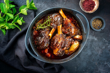 Modern style traditional braised slow cooked lamb shank in red wine sauce with shallots and carrots offered as top view in a design stewpot - obrazy, fototapety, plakaty