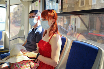 Young red-haired woman wearing protective face mask sits next to man in public transport on sunny summer day. Safety requirements during pandemic