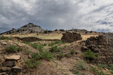 Fototapeta na wymiar Few levels of ruined stone walls in the foreground against a high hill with medieval walls and towers and a dramatic grey sky. These are monuments of the Great Genoese civilization in Crimea.