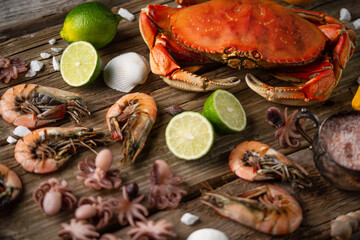Top view of tiger shrimps, cooked crab and baby octopuses with lime, lemon and seashells on rustic wooden background. Concept of delicious food from the sea or ocean.