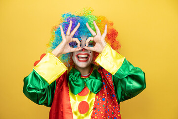 Clown standing over yellow insolated yellow background doing ok gesture shocked with smiling face, eye looking through fingers