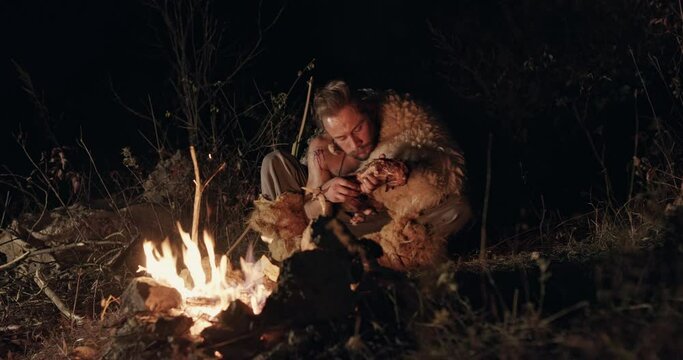 Hungry wild man eating near campfire. Low angle of hungry male barbarian eating meat while sitting near fire at night in countryside