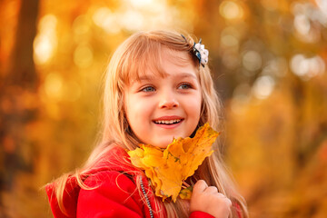 Naklejka na ściany i meble Outdoor portrait of happy blonde child girl in a red jacket holding yellow leaves. Little girl walking in the autumn park or forest. Warm sunny weather. Fall concept