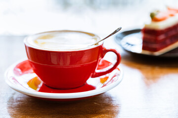 Close up of a red cup of coffee on a wooden table. In the background is a black plate with strawberry cake