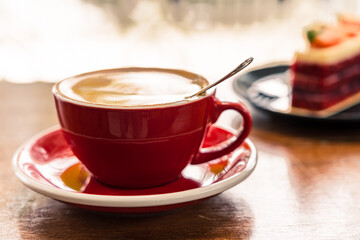 Close up of a red cup of coffee on a wooden table. In the background is a black plate with strawberry cake