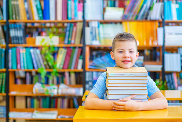 A boy sitting in the library hugs a stack of books lying in front of him on the table with a smile looking at the camera