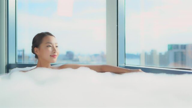 Young asian woman taking a hot bath with white foam covering her body, admiring the city view and relaxing.
