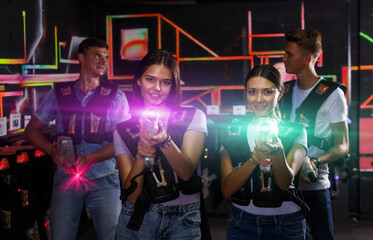Obraz na płótnie Canvas Two girls standing and holding guns during laser tag game with men in dark labyrinth