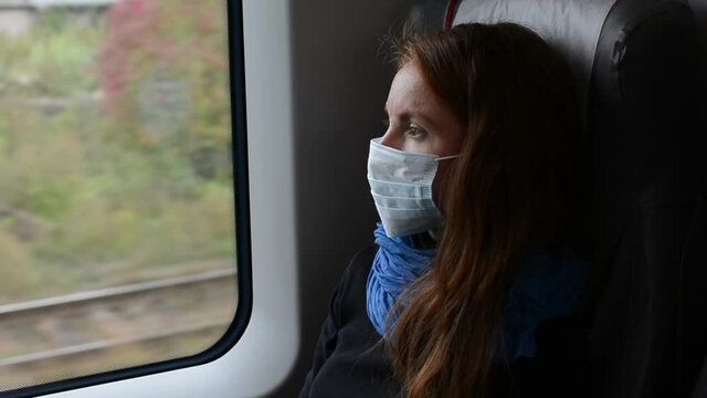 Young lonely woman looks through the window in a medical mask in train during the coronavirus epidemic, close up portrait. Coronavirus prevention, social distancing in public transport for passengers.