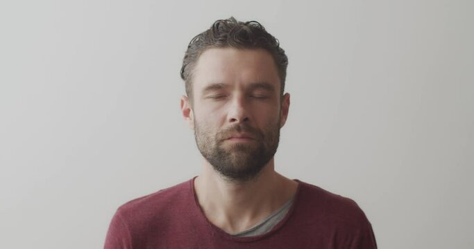 Close up front view of male face doing yoga exercises indoors on white background. Bearded man with closed eyes practicing pranayama natural light slow motion. Healthy body health care concept
