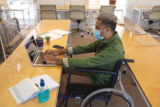 Disable man wearing face mask using laptop while sitting on his wheelchair at office