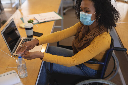 Disable woman wearing face mask sanitizing her hands while sitting on wheelchair at office