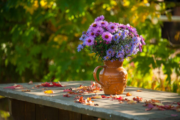 Bouquet of fresh purple autumn flowers in vintage vase on wooden table in outdoor garden patio with some autumn leaves on and sunlit background. Copypaste. Concepts: cosy, bright, natural