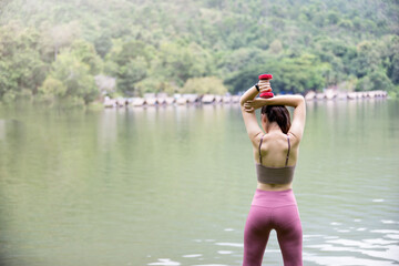 Rear view of female fitness instructor exercising with small dumbbell on green grass near lake in...