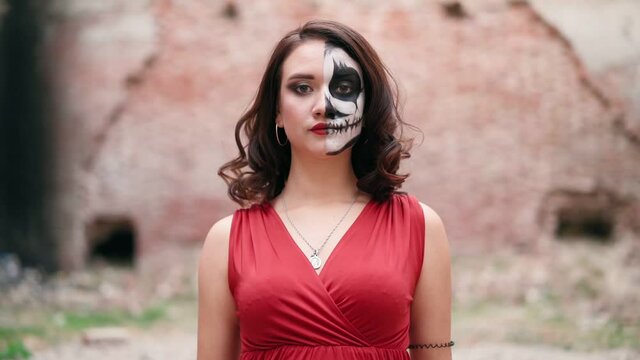 Portrait beautiful woman in a red evening dress and awesome Halloween makeup goes against the backdrop of an abandoned building. Greasepaint for the Holiday on October 31. Slow motion