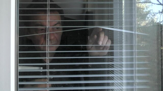 A man is observing with binoculars through the blinds in the window.