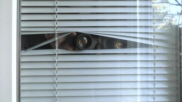 A man is observing with binoculars through the blinds in the window.